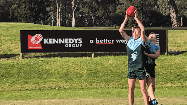 two children playing AFL on a geelong ground with kennedys Group sponsorship signage